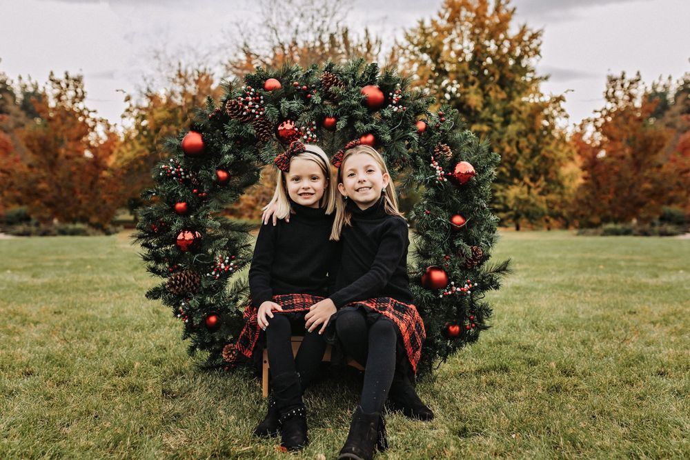 Matching Christmas outfits seated in front of large decorated wreath outdoors.