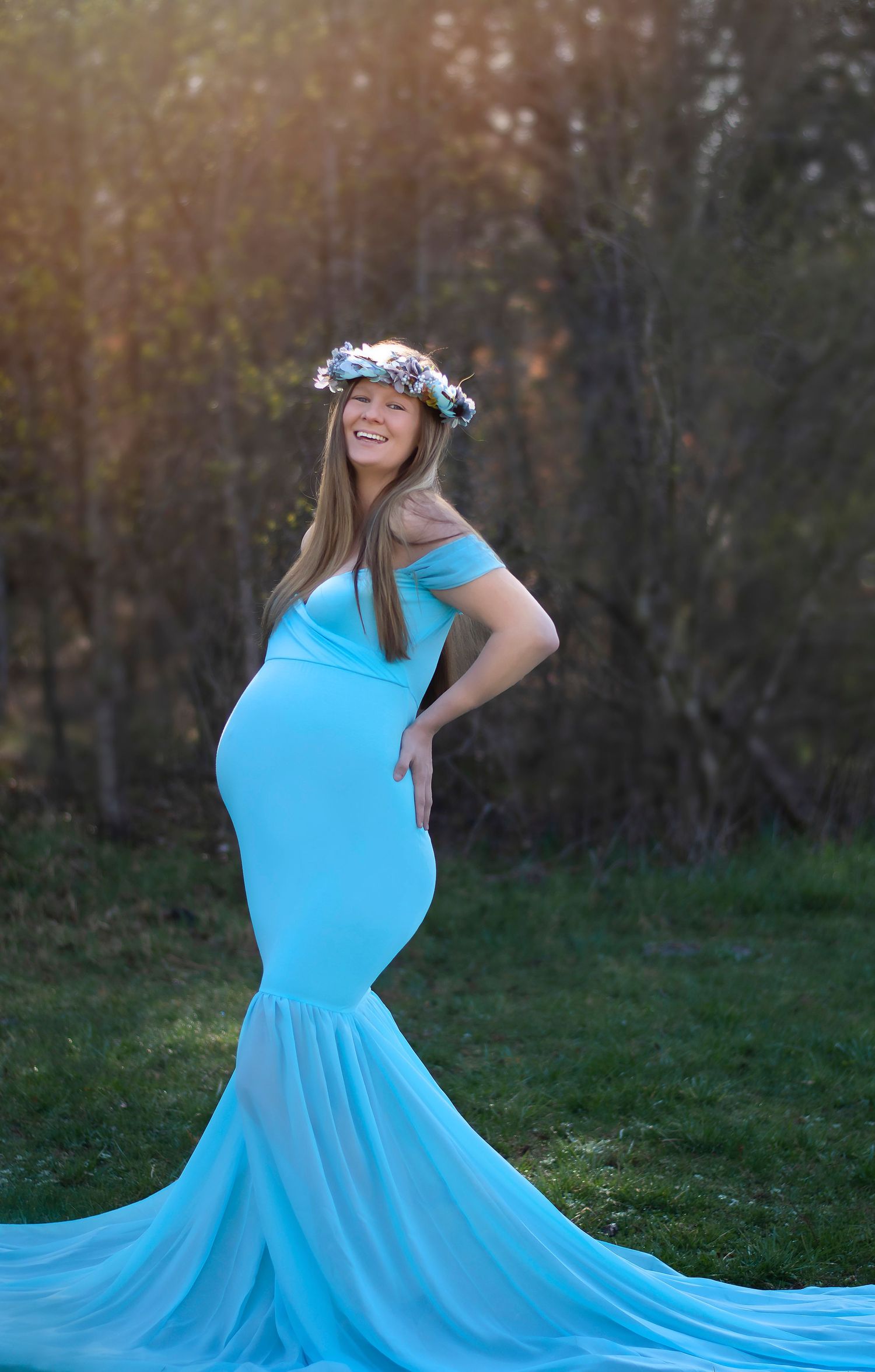 Maternity portrait in bright blue gown with flower crown against forest backdrop at sunset.