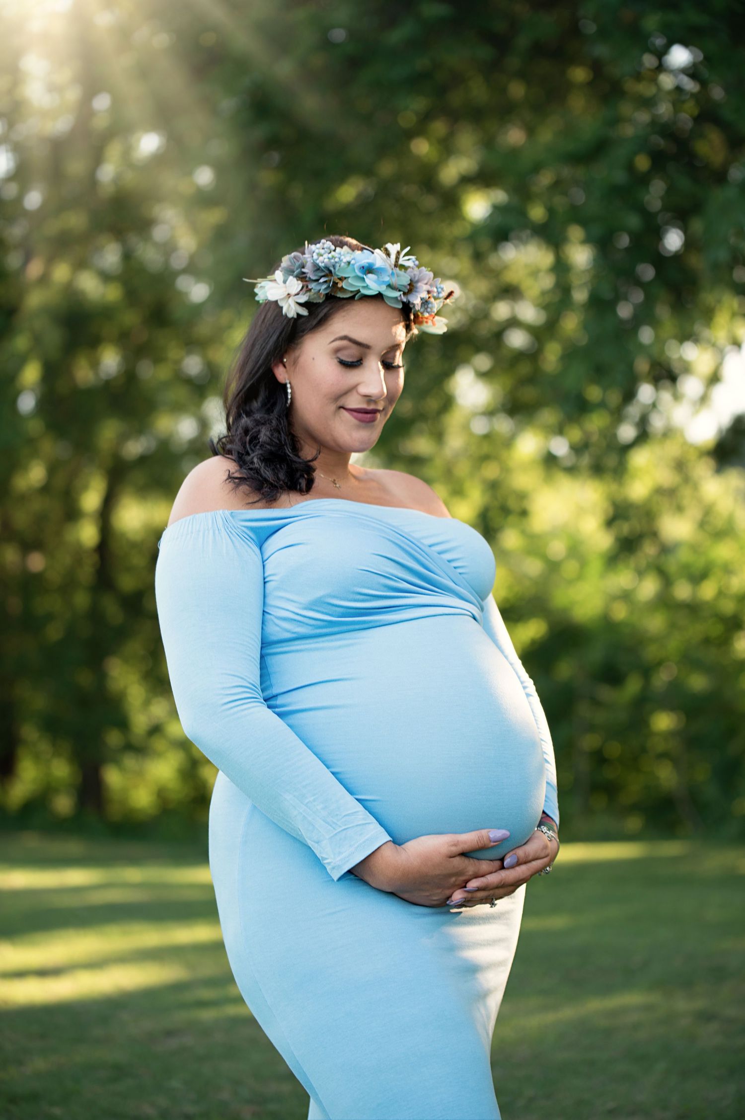 Expectant mother in light blue off-shoulder dress wearing floral crown poses in sunlit garden setting.