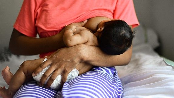Woman breastfeeding a dark-haired baby in the football position sitting on a bed