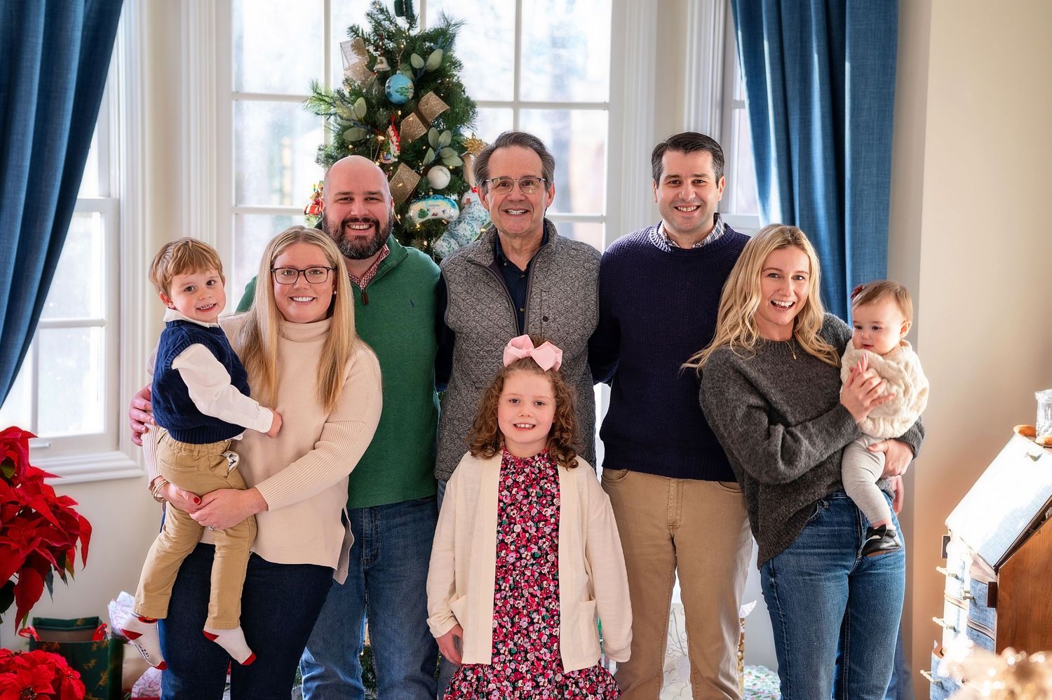 A cheerful family gathering in front of a decorated Christmas tree in a living room with blue curtains.