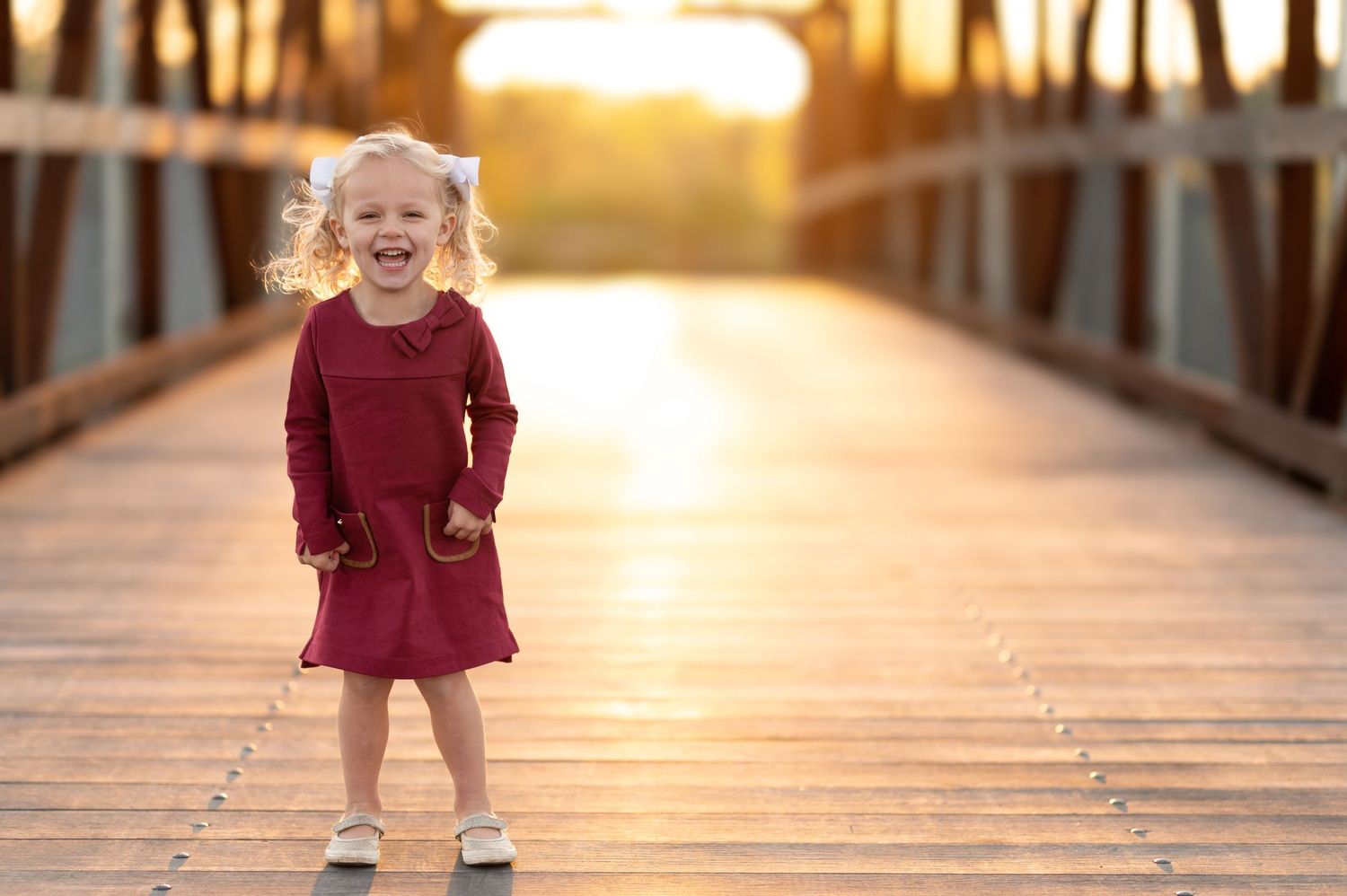 Young child in burgundy dress laughs joyfully on wooden bridge at sunset.
