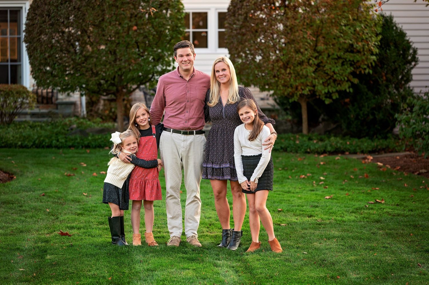 A family stands together on a green lawn with autumn trees in the background during a photo session.