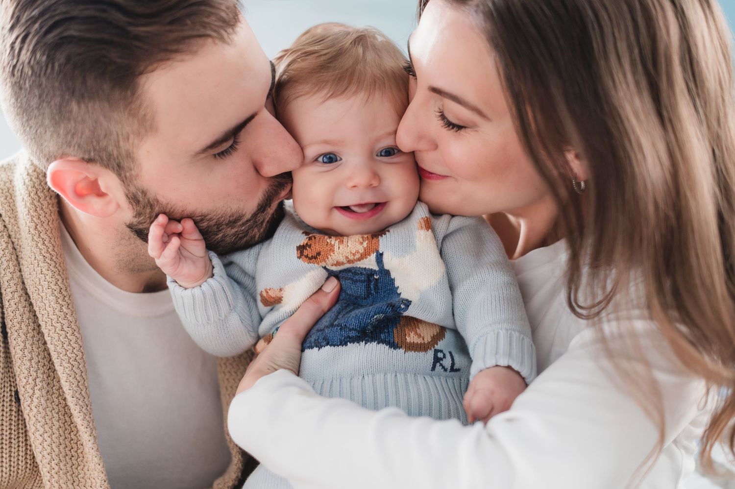 A tender family moment with a baby in denim overalls being kissed by parents.