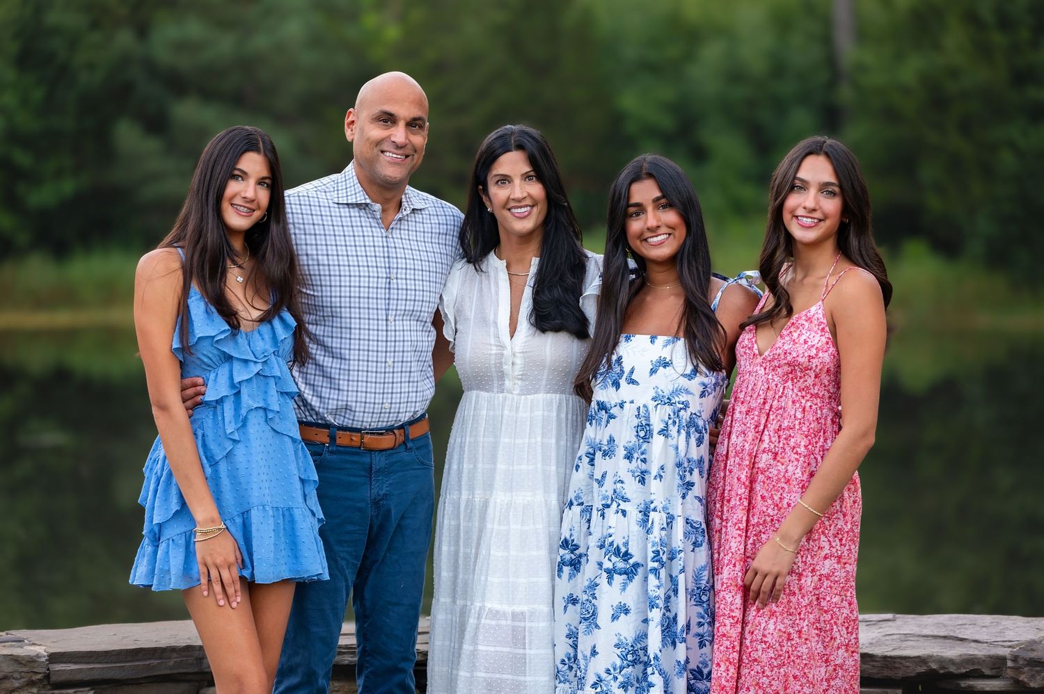 A family group in coordinated summer dresses and business casual attire poses outdoors near a wooden railing.