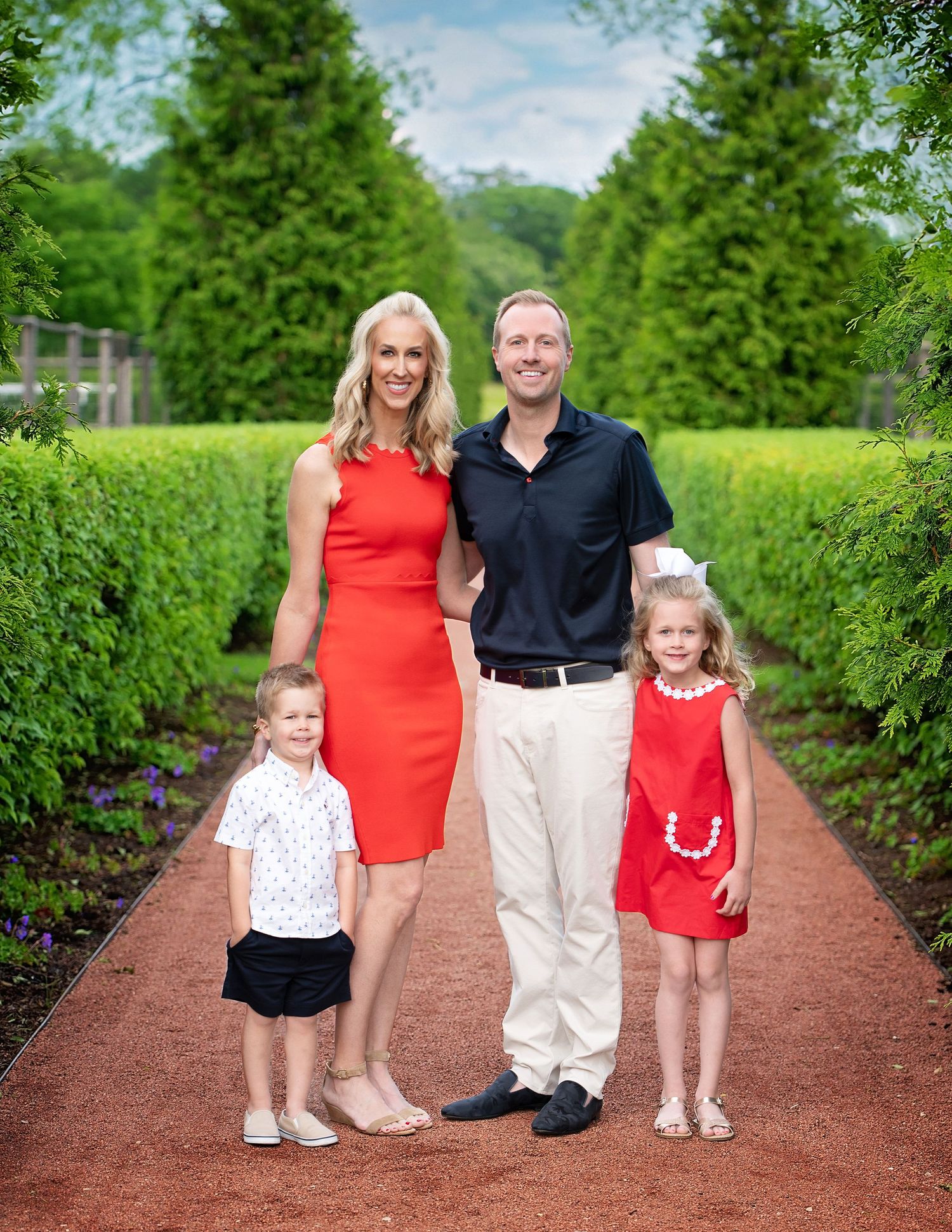 A family of four in coordinating red and white outfits poses in a formal garden with manicured hedges.