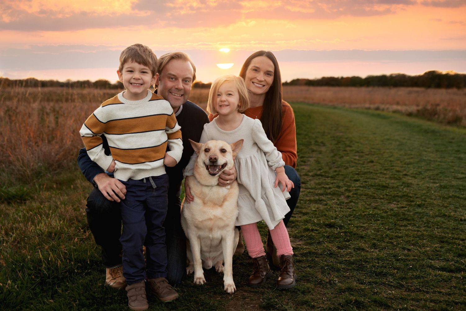 Family and their golden retriever pose together at sunset in a grassy field.