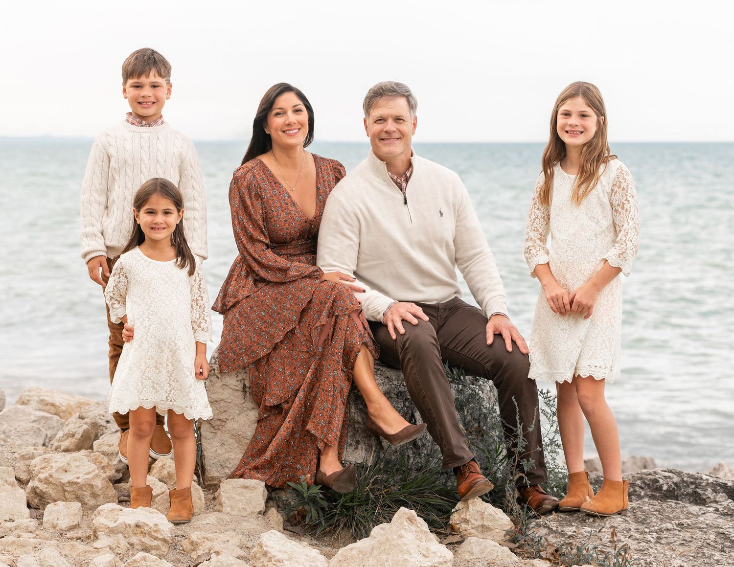 A family in neutral clothing poses together on rocky beach with ocean backdrop.