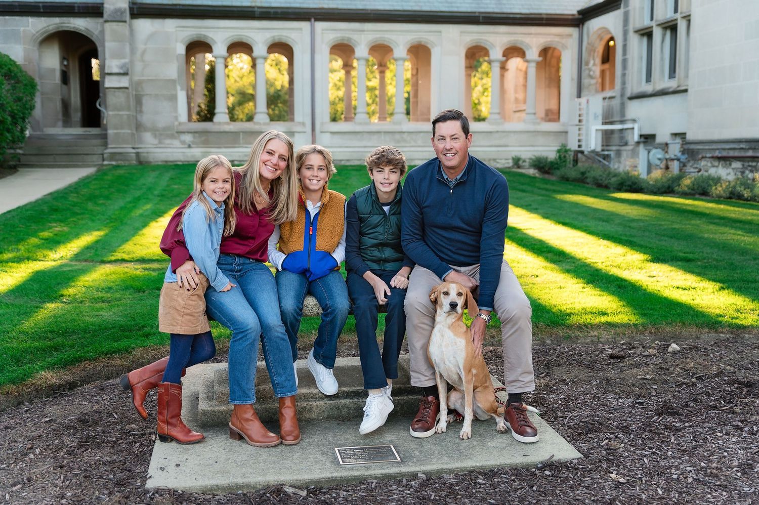 A family and their dog sit together on stone steps with arched architecture and sunlit grass in background.