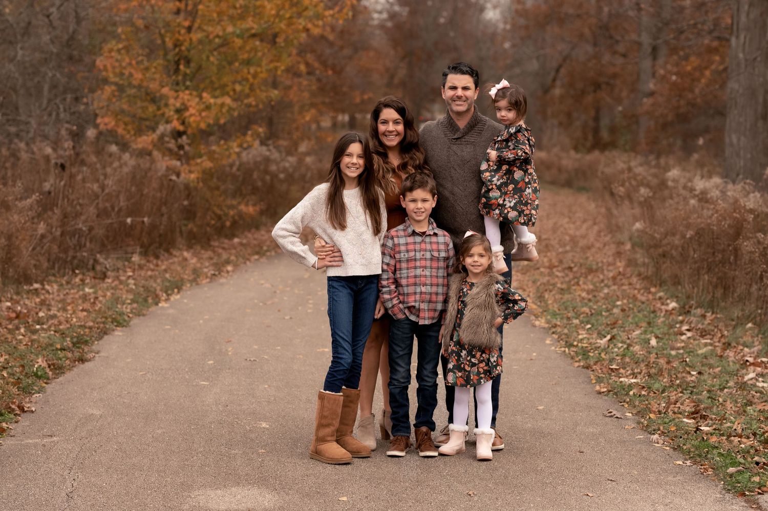 Family portrait on a tree-lined autumn path with fallen leaves.