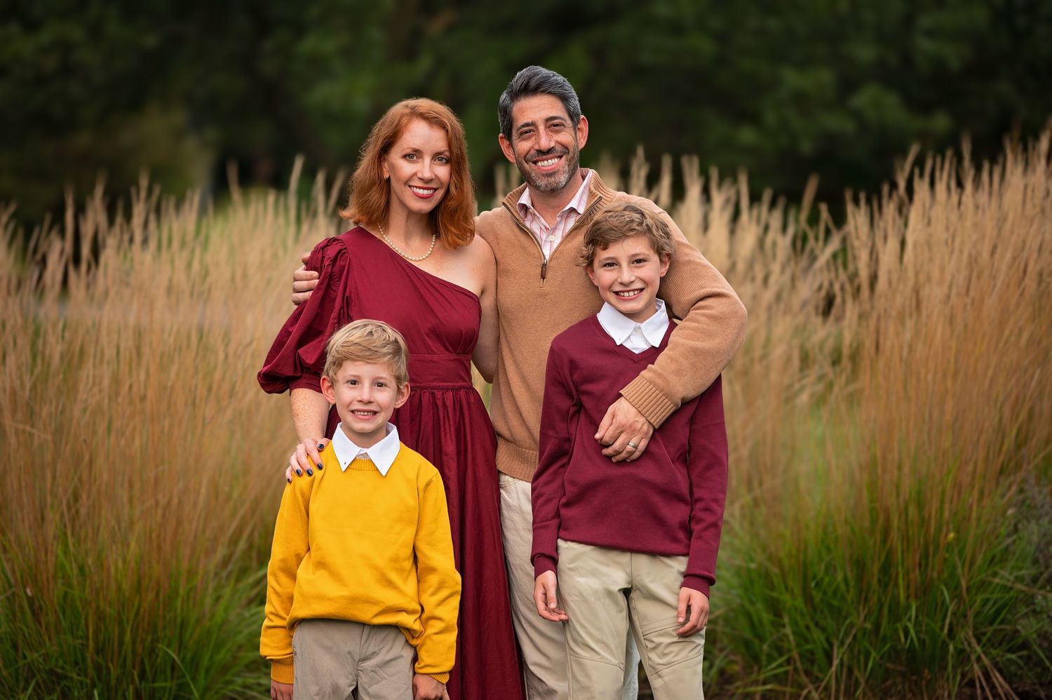 A family of four poses together in front of tall autumn grass wearing coordinated burgundy and yellow outfits.