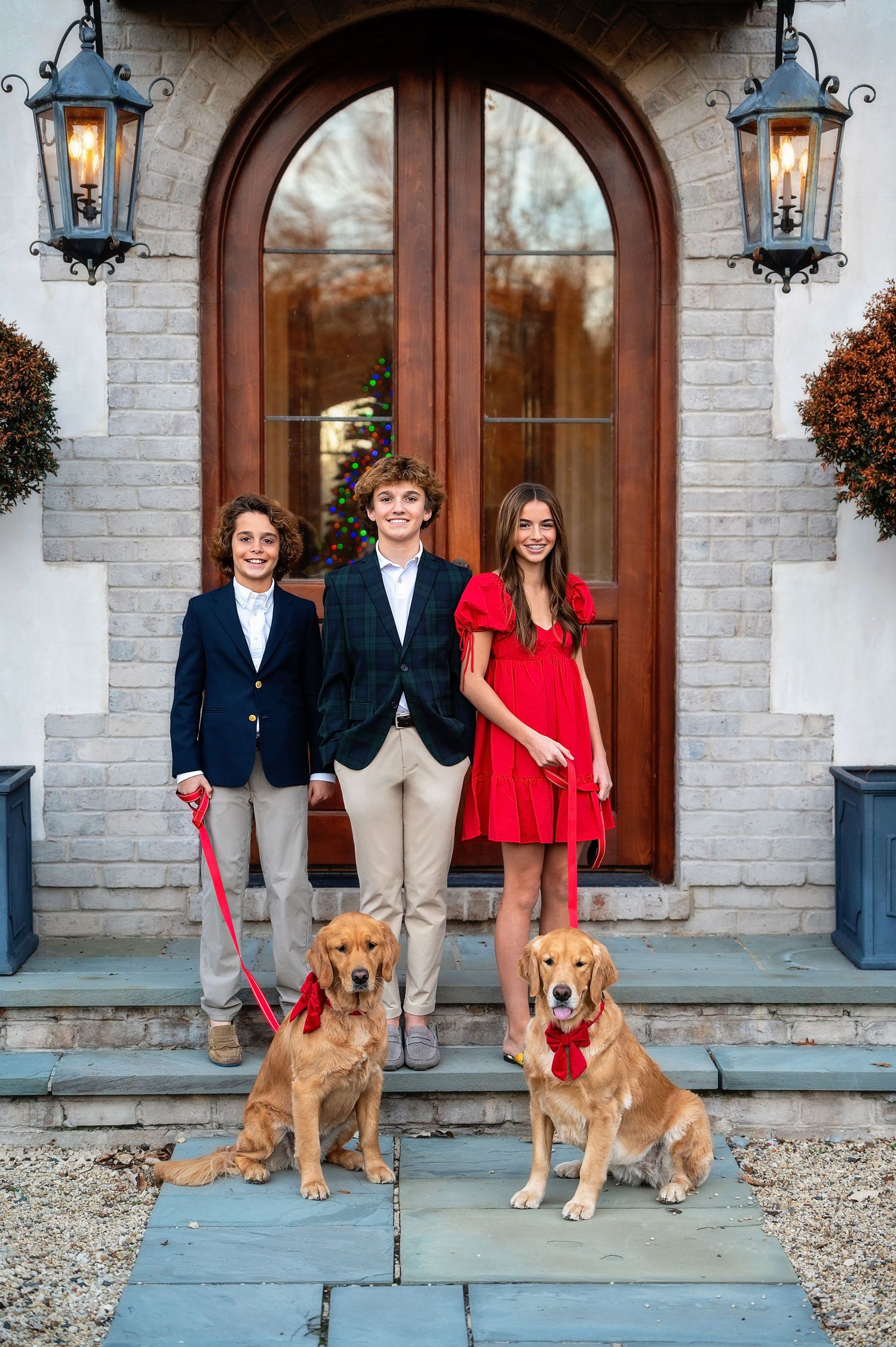 Three people and two golden retrievers pose on stone steps in front of an arched wooden doorway with lanterns.