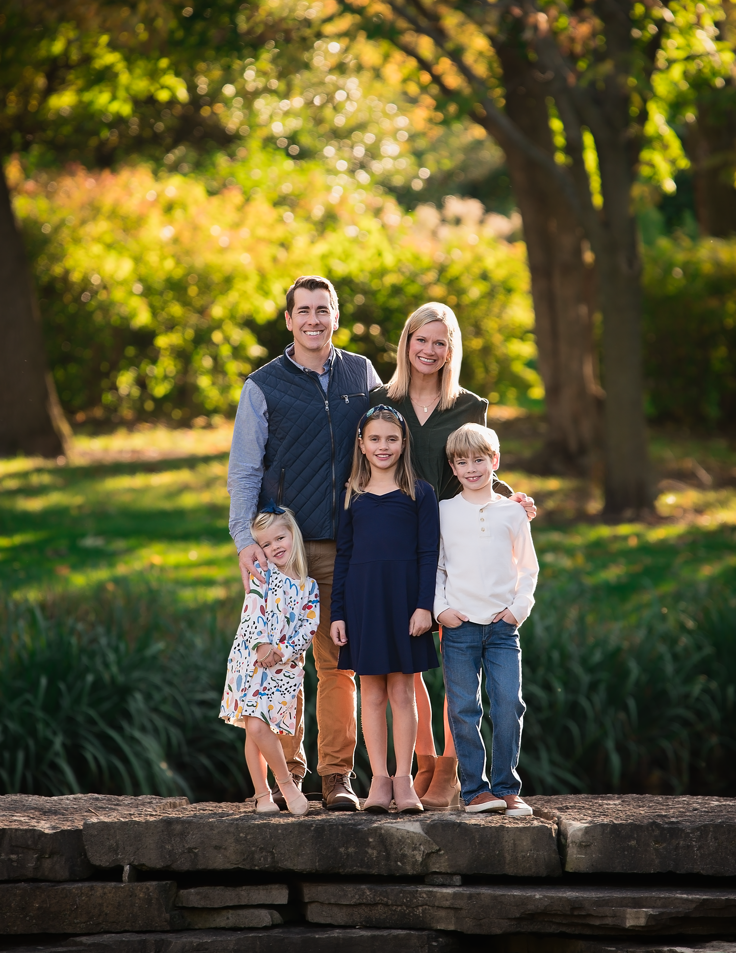 A family poses together on stone ledge with autumn foliage in background during outdoor photo session.