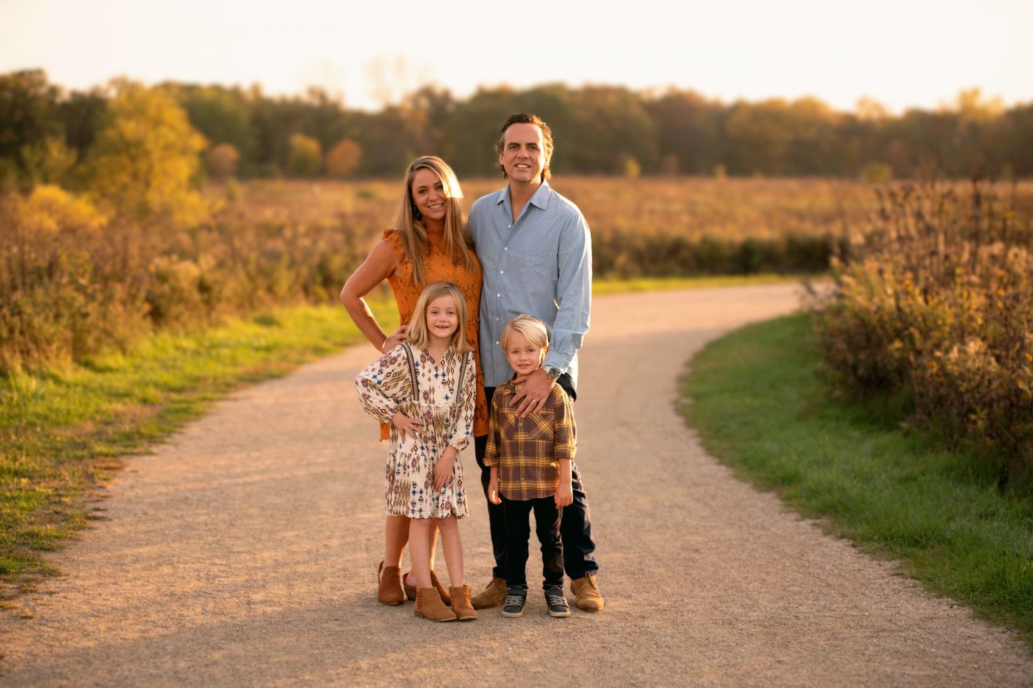 Family of four poses on dirt path during golden hour surrounded by autumn fields.