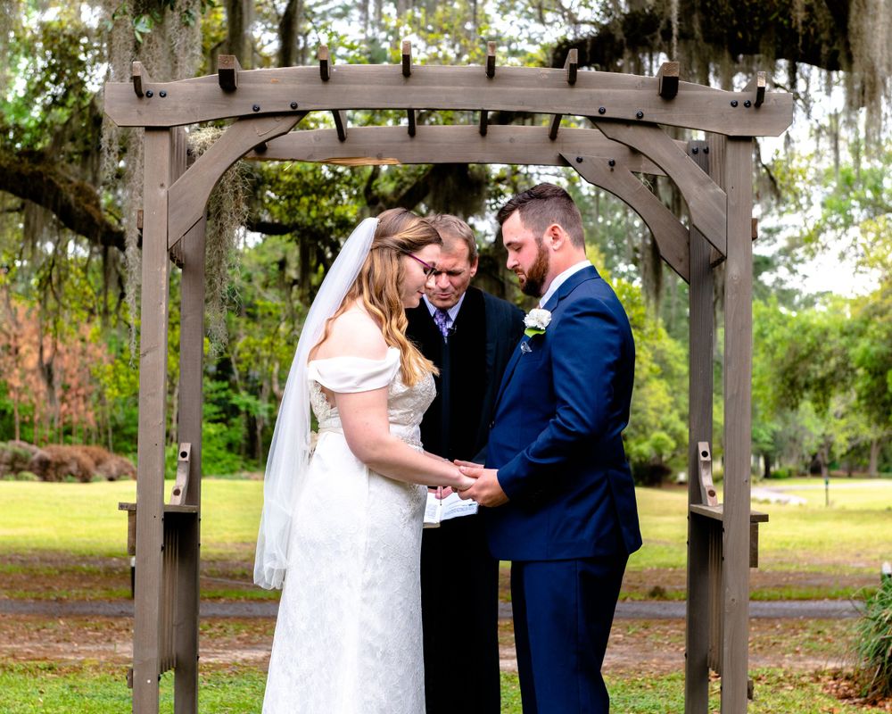 Wedding ceremony under rustic wooden pergola with hanging Spanish moss in garden setting.