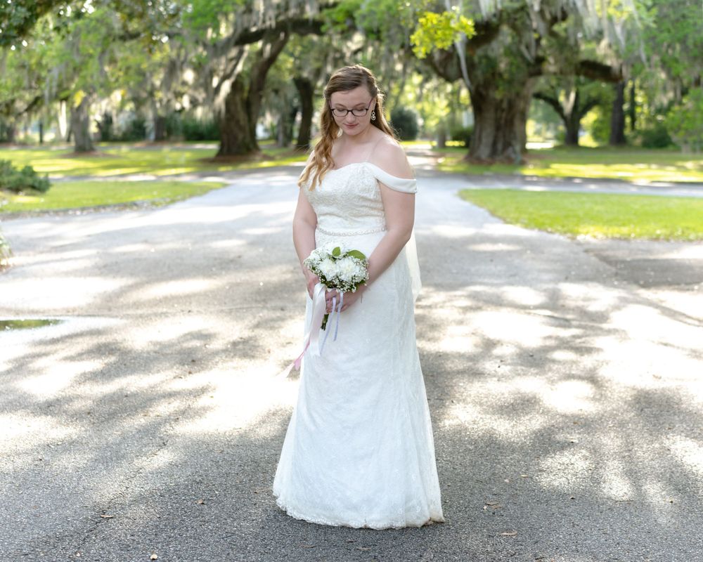 Bride in off-shoulder lace gown holds bouquet while standing on sunlit garden path.