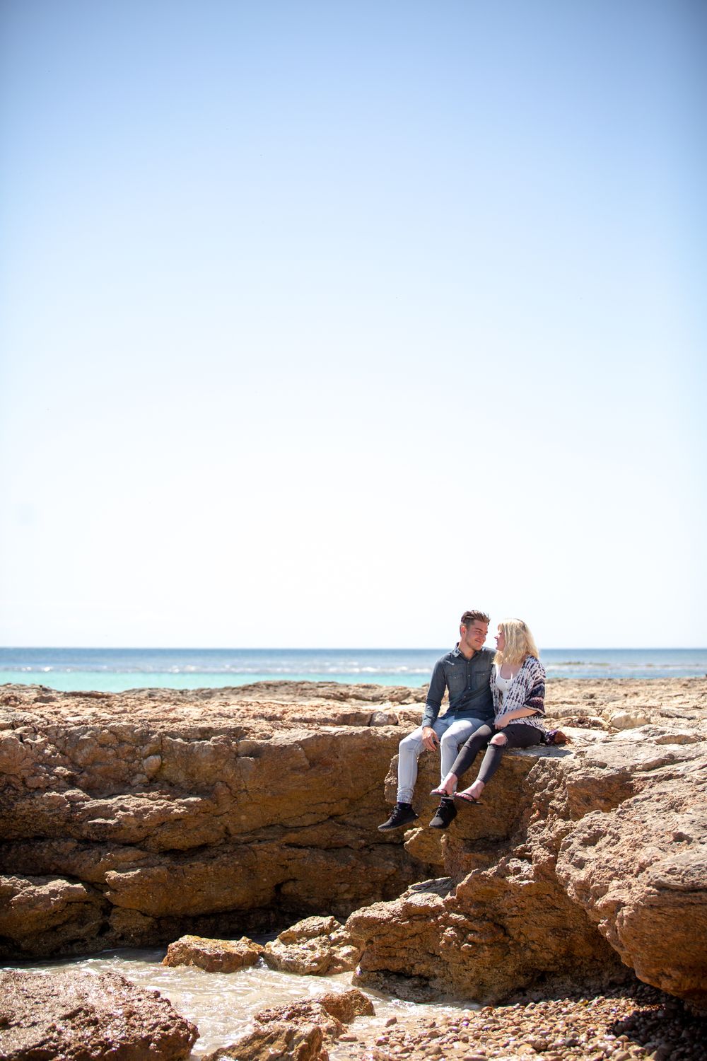 newly engaged couple sitting on the cliff with water surrounding them smiling at each other
