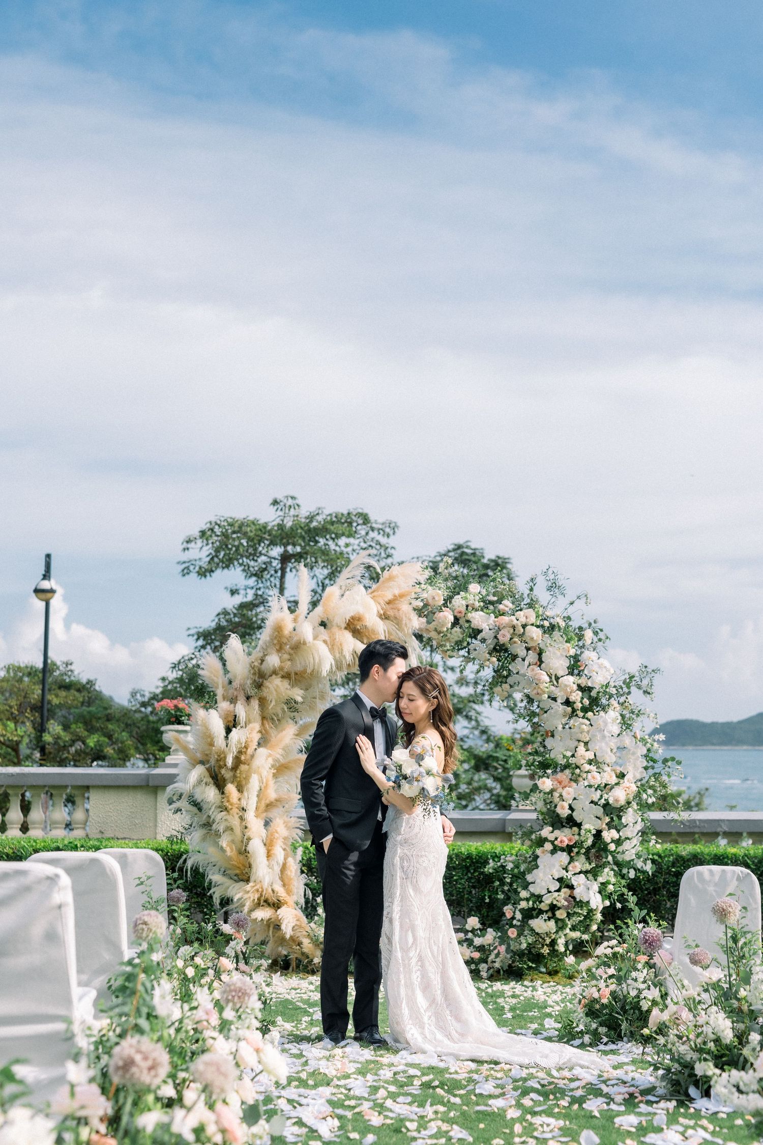 Romantic wedding couple embracing under a floral arch with scenic ocean view.