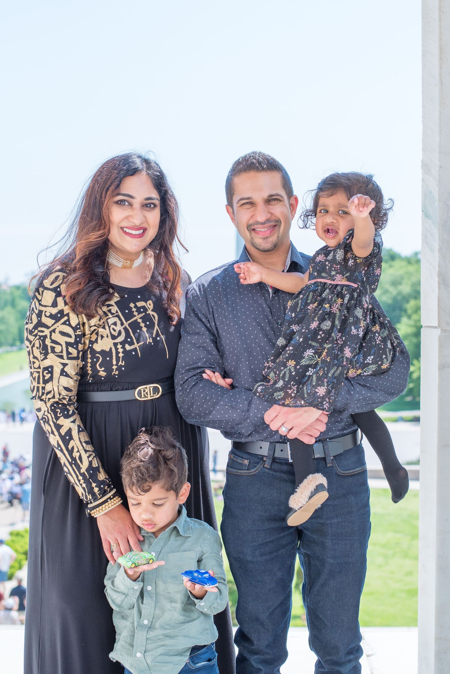 family of four look at camera with Washington Monument behind them, daughter reaches her hand towards camera