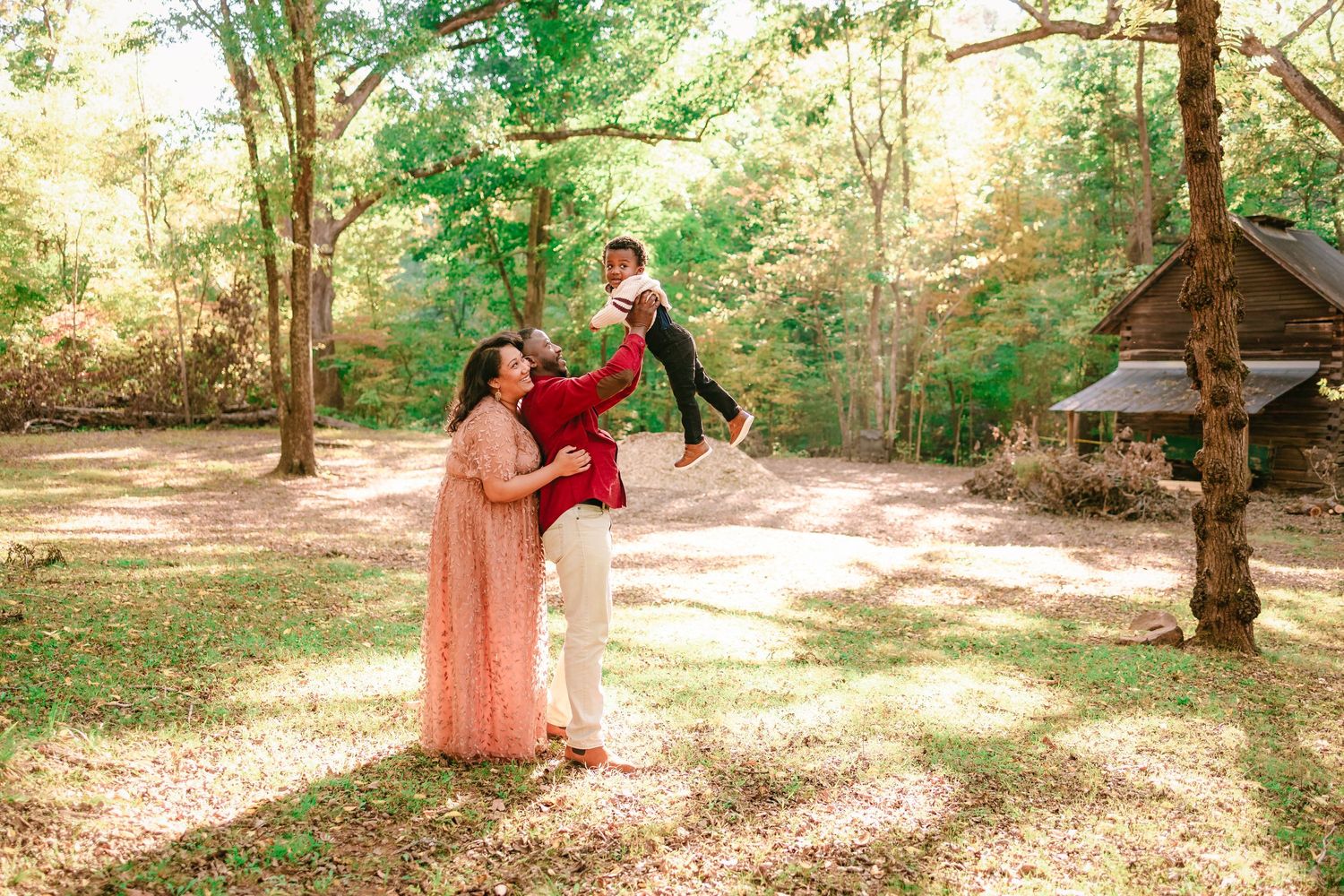 Family in traditional attire playing outdoors, parents lifting child in sunlit forest clearing.