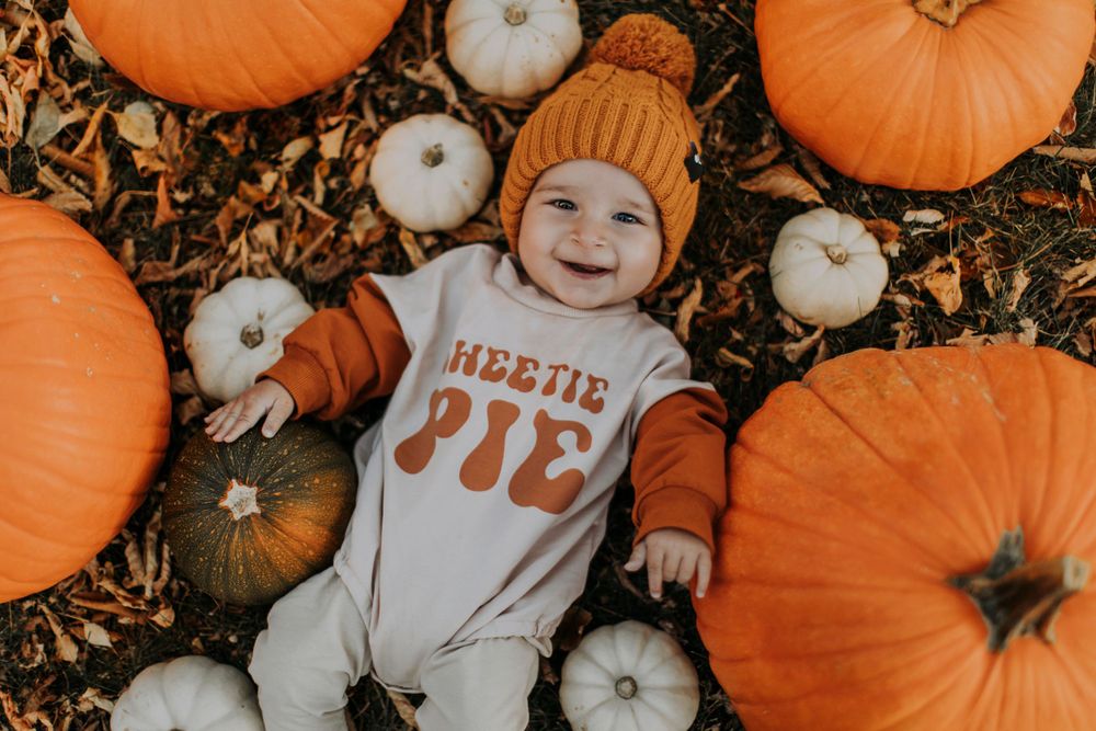 A baby wearing an orange beanie lies surrounded by orange and white pumpkins on autumn leaves.