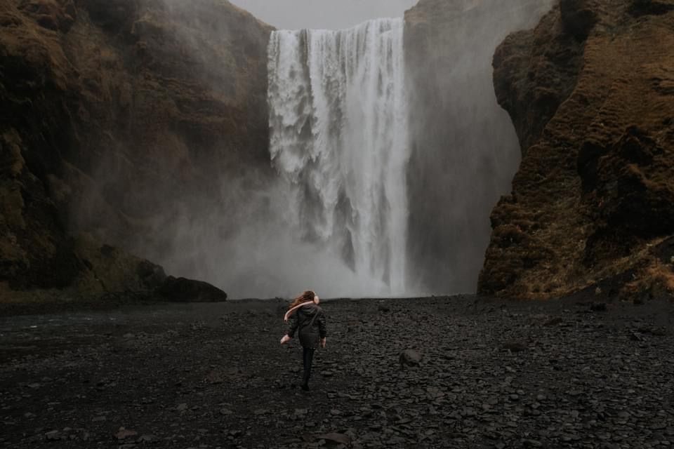 A distant figure stands before a massive waterfall surrounded by dark rocky terrain.