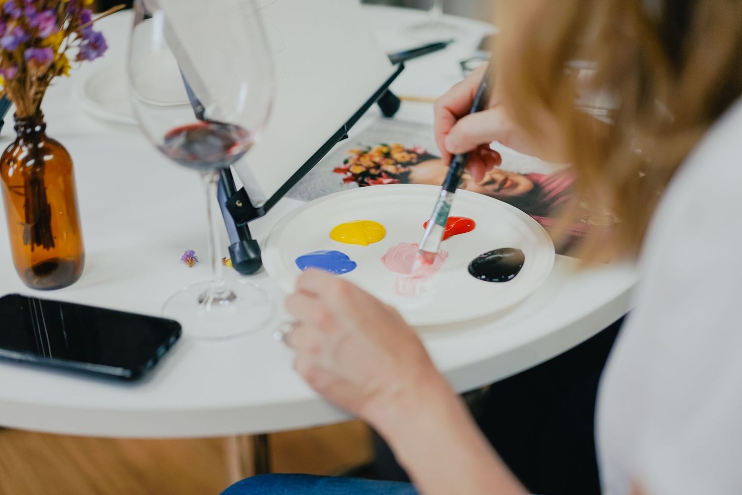 Paint palette with colorful paints and brushes on white table with flowers in amber vase.