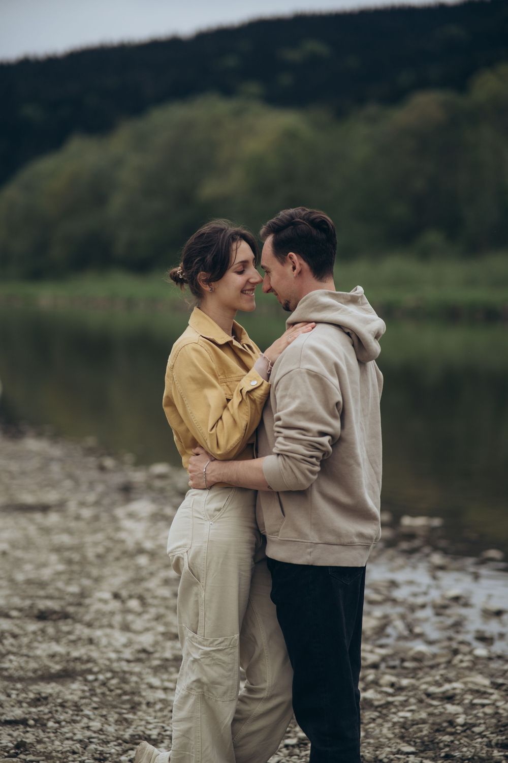 Couple sharing a tender moment by a peaceful lake with forest backdrop.
