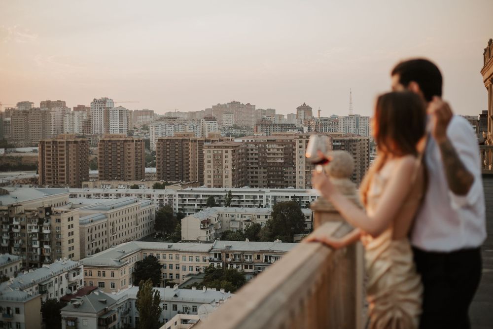 Couple enjoying sunset view of city skyline from rooftop balcony with tall buildings in background.
