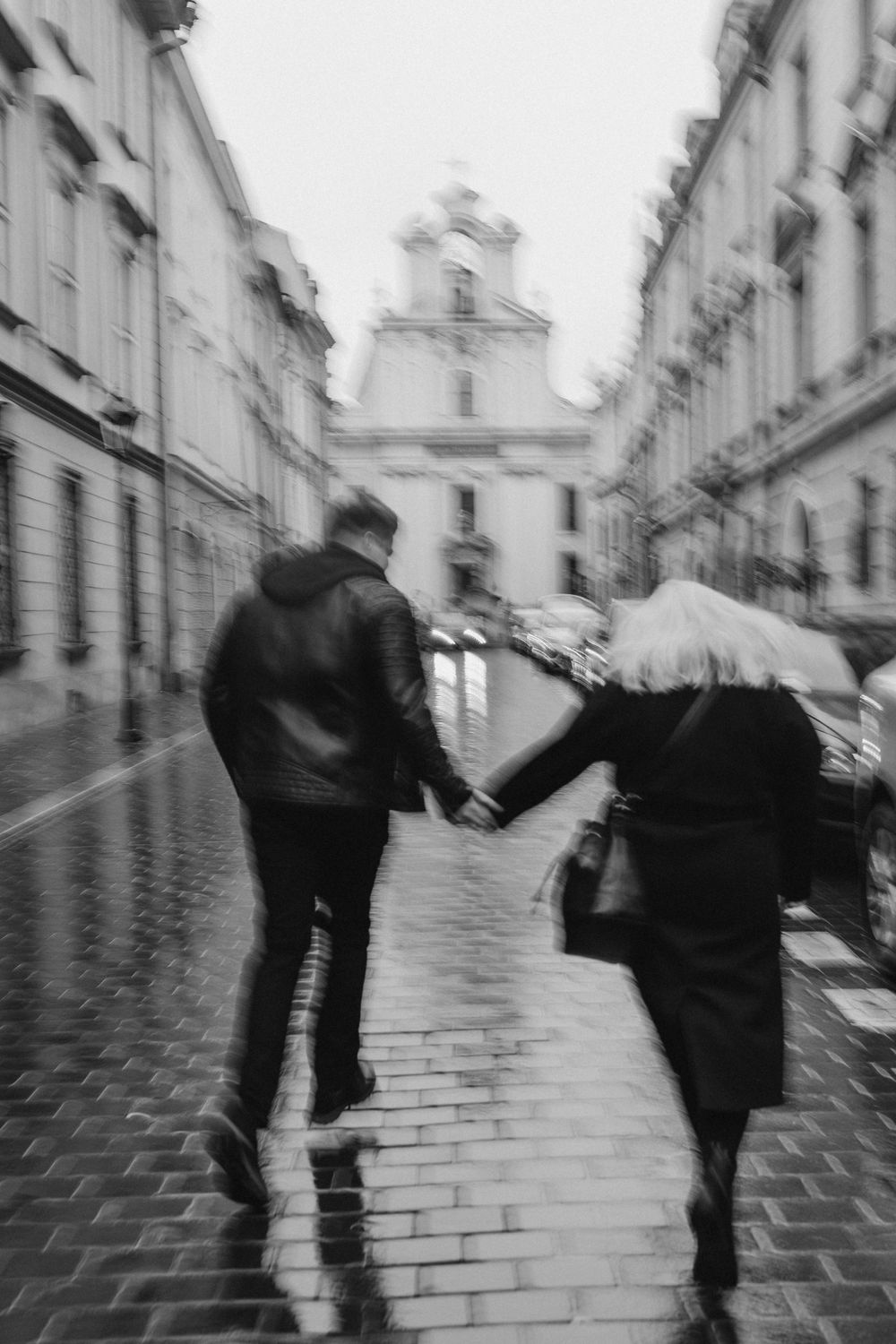 Blurred motion shot of couple running in rain on cobblestone street near historic buildings.