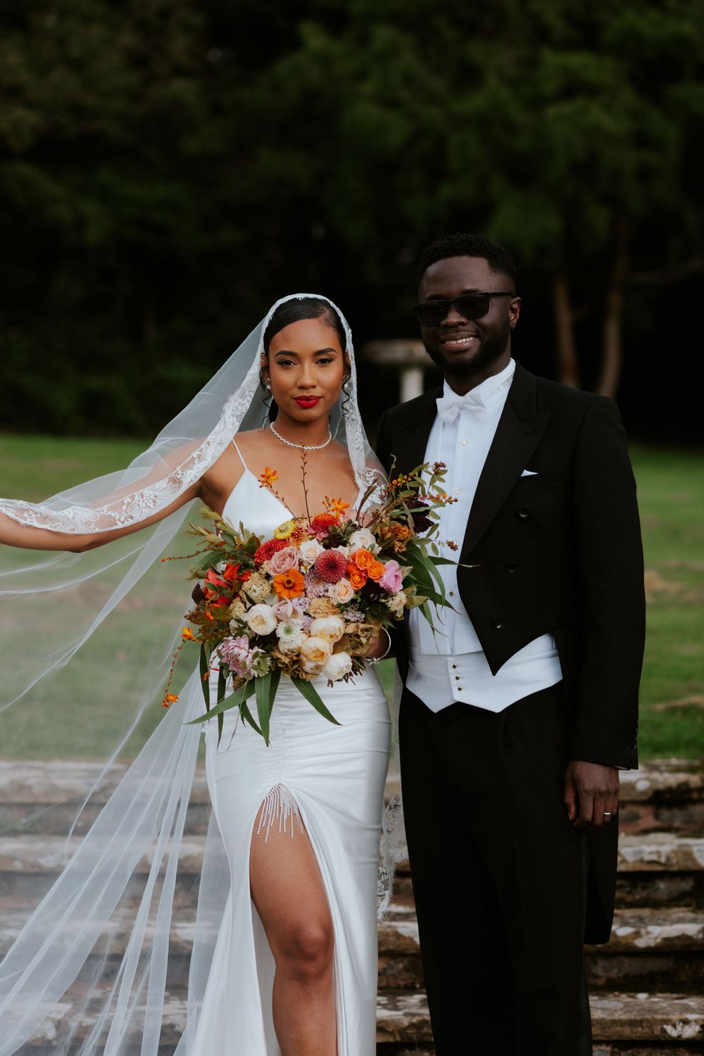 Elegant wedding portrait with flowing veil and vibrant autumn-colored bouquet against garden backdrop.