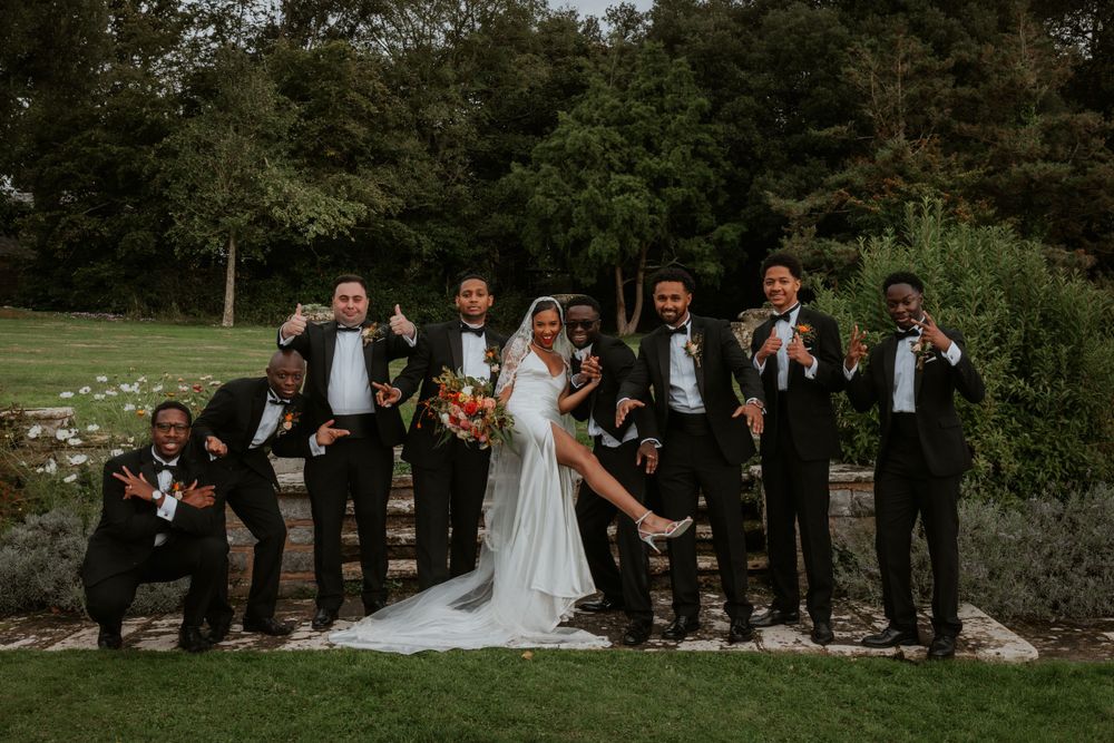 Wedding party in formal black attire poses playfully on garden steps.
