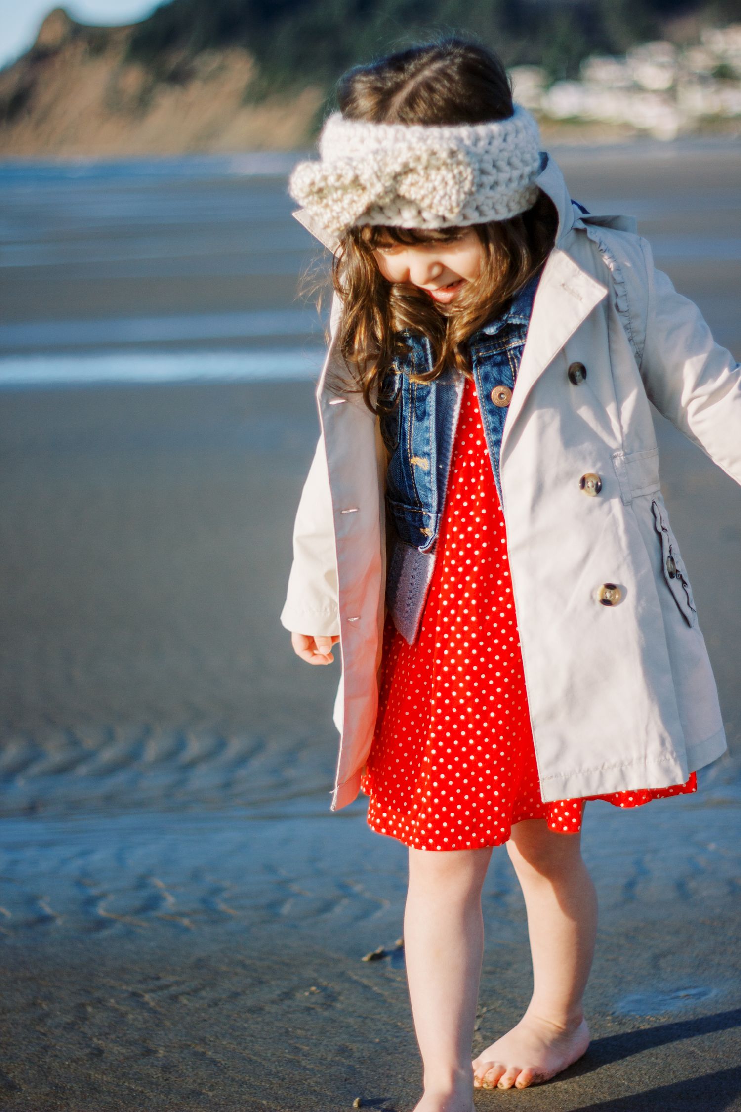 Child in white coat and red polka dot dress playing barefoot on a beach.