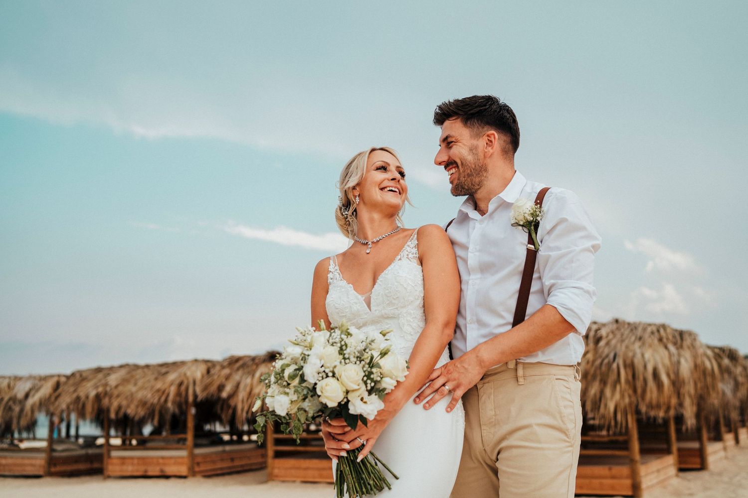 A joyful beachside wedding portrait with rustic thatched umbrellas in the background.