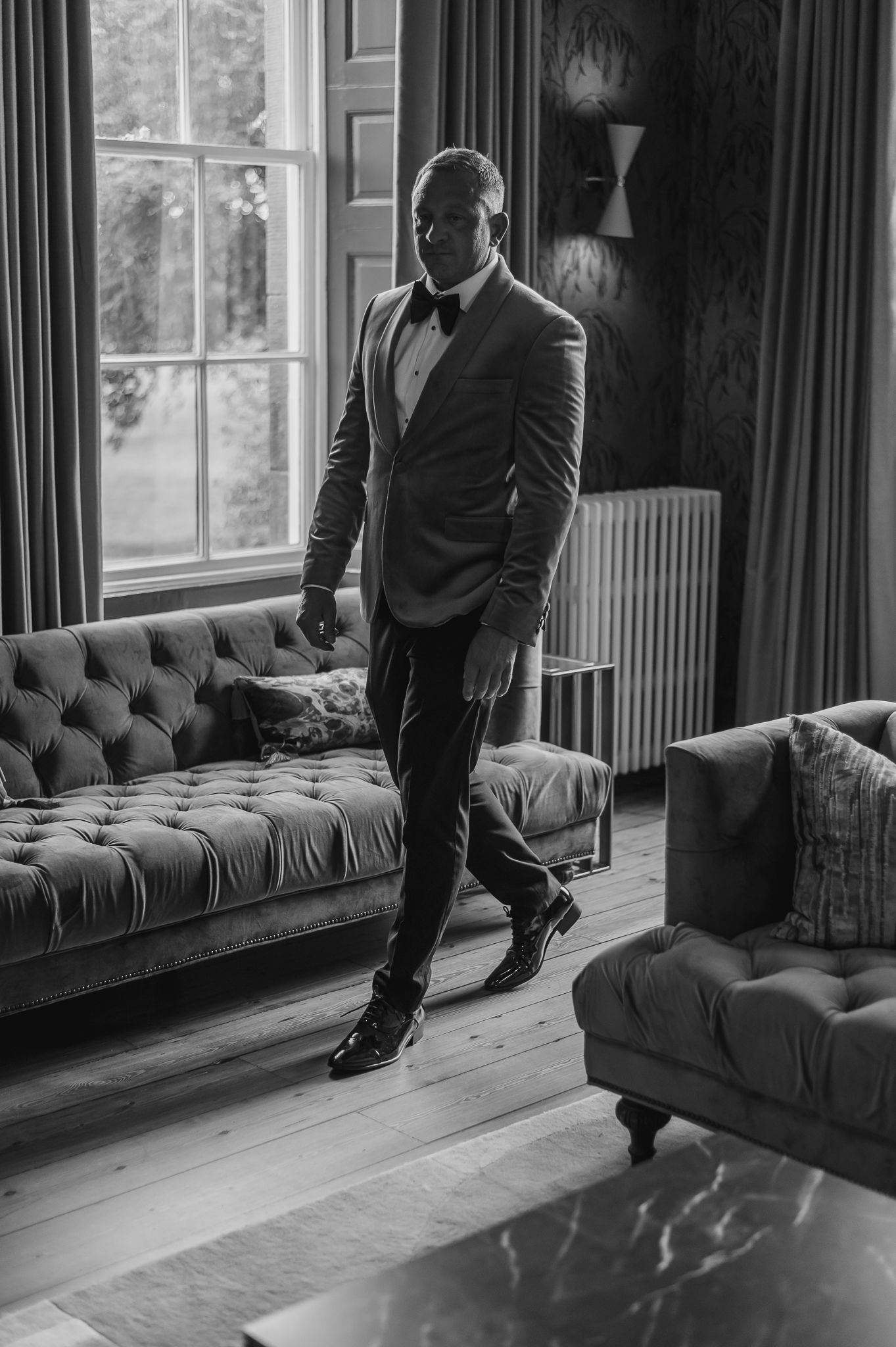 Elegant black and white photo of someone in formal attire standing by a tufted sofa near a window.