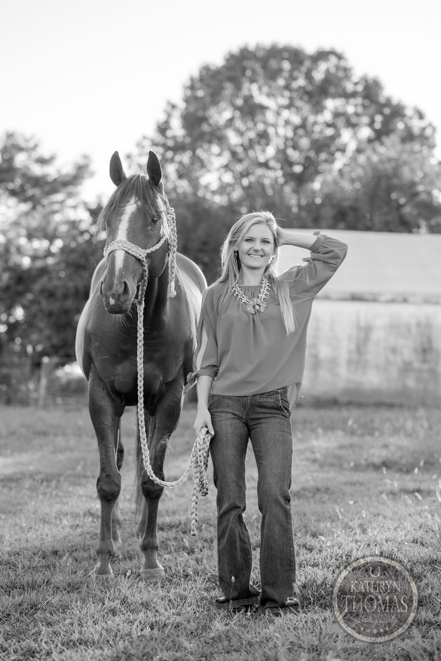 A black and white photo showing a person posing next to a beautiful horse in a pastoral countryside setting.