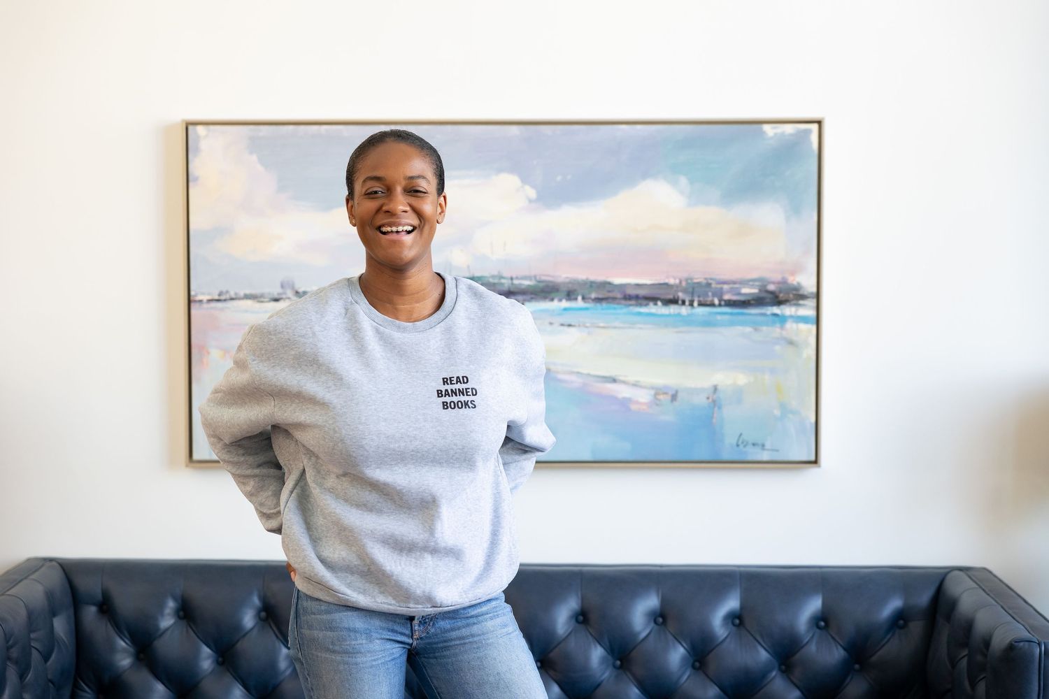 Smiling woman in gray sweatshirt standing in front of a large abstract painting on a couch.