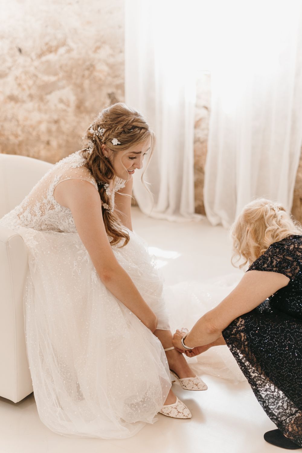 Bride in lace wedding gown smiles as woman helps fasten her white bridal shoes.