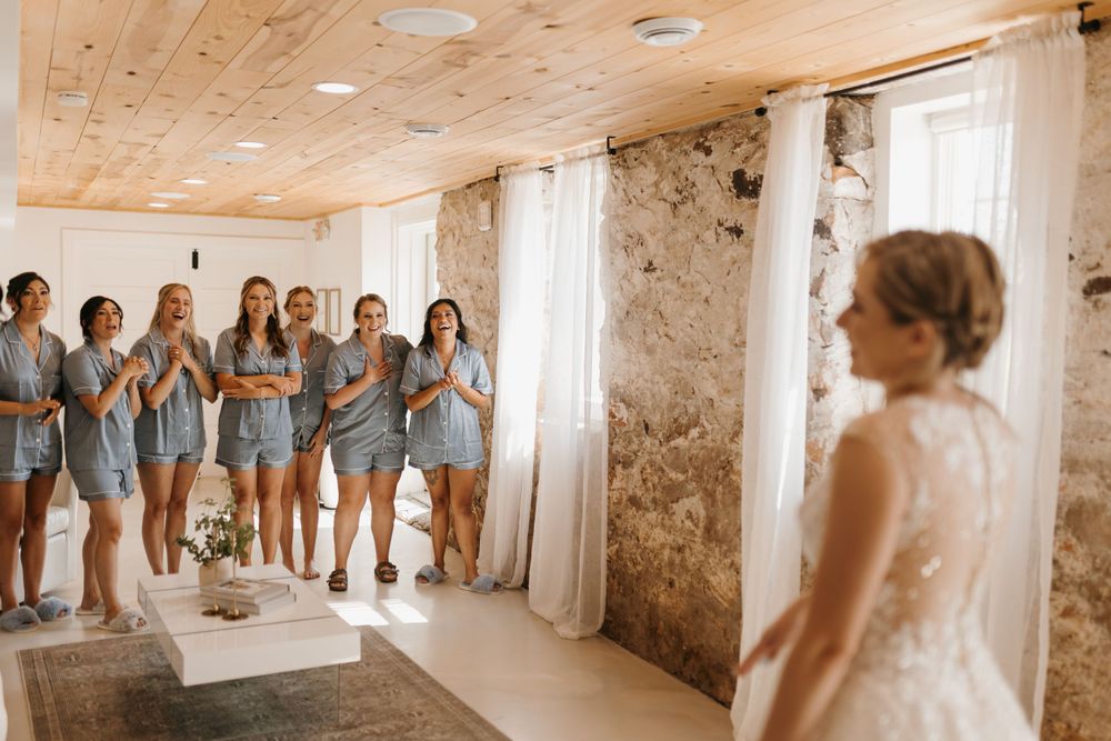 Bridesmaids in blue matching sets clap and react emotionally as bride in lace wedding gown turns to reveal her look.