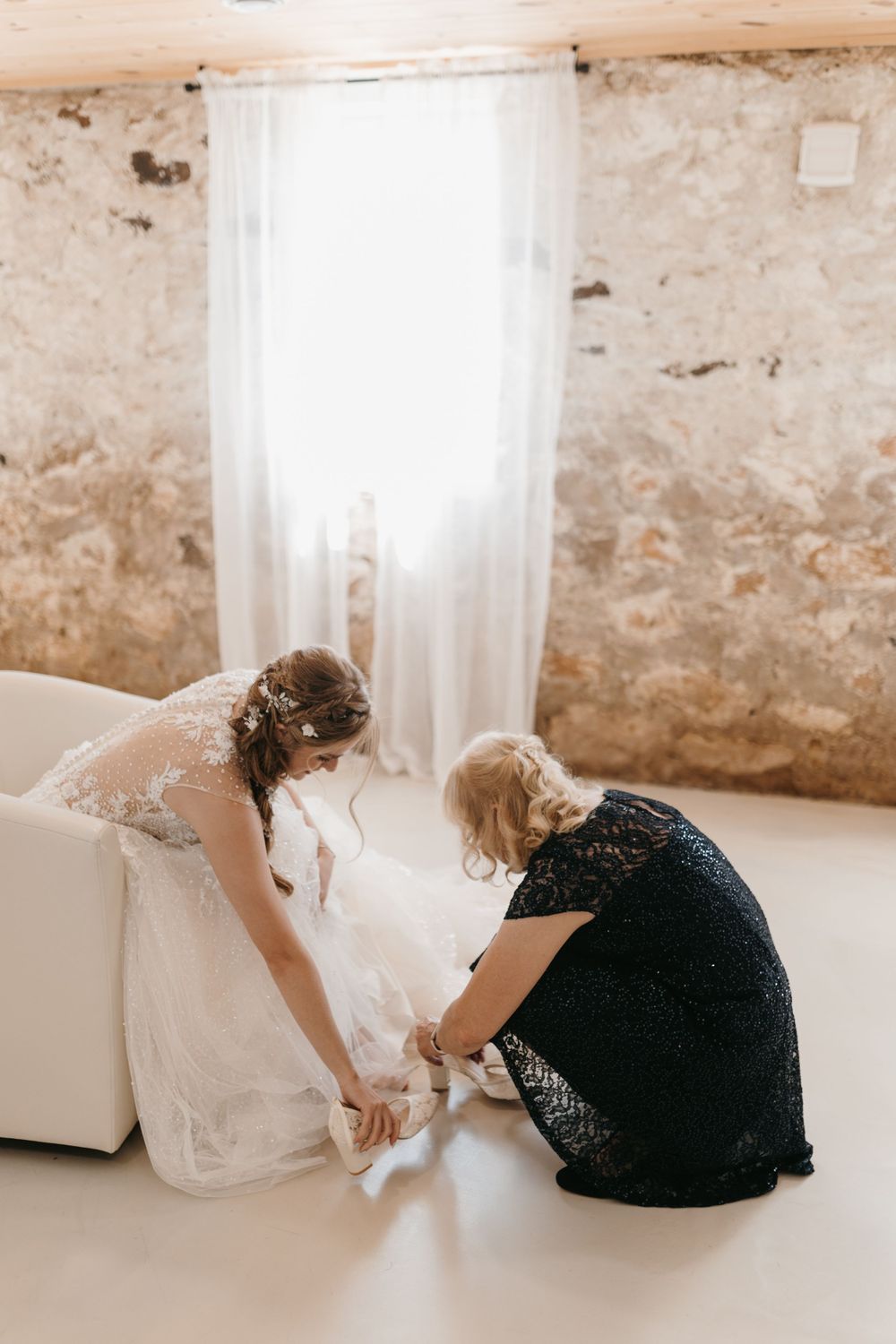 Woman in black lace dress helps bride put on wedding shoes in bright rustic venue.