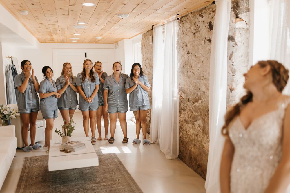 Bridesmaids in matching blue pajamas react with joy seeing bride in stunning lace gown during emotional first look.