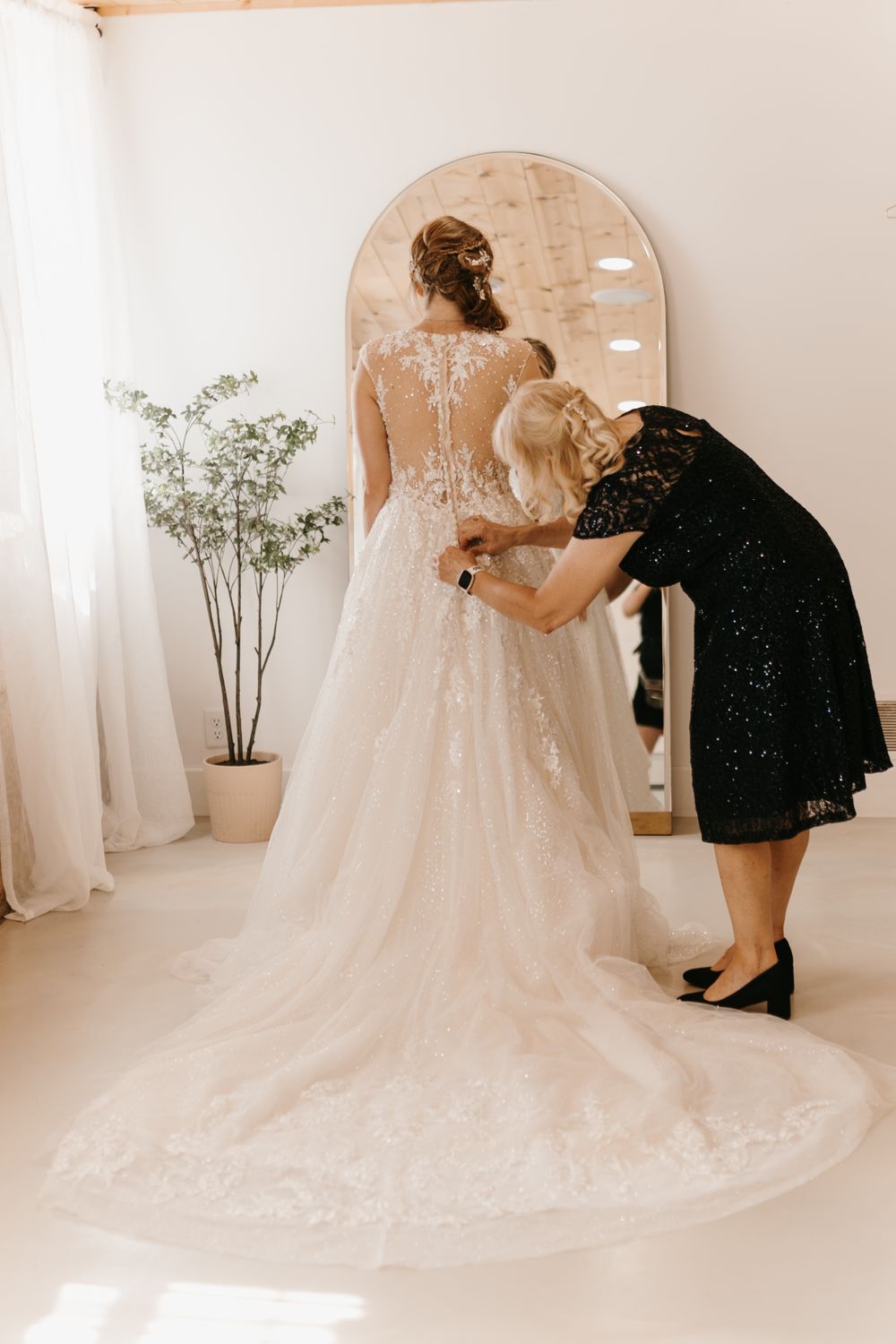Woman in black lace dress buttons up back of brides elegant lace wedding gown.