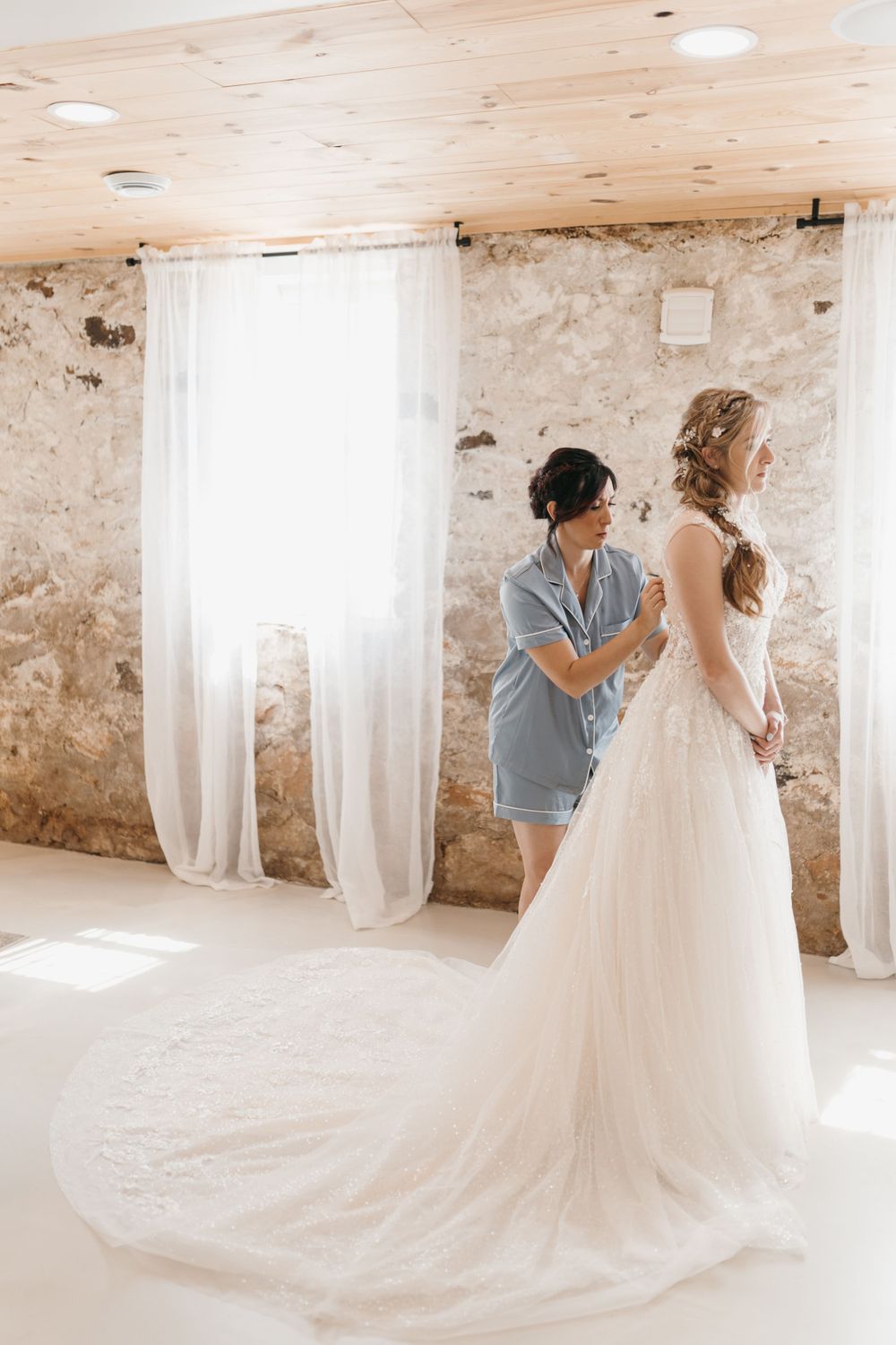 Bridesmaid in blue pajamas helping bride button up elegant lace wedding gown with long train in bright rustic venue.