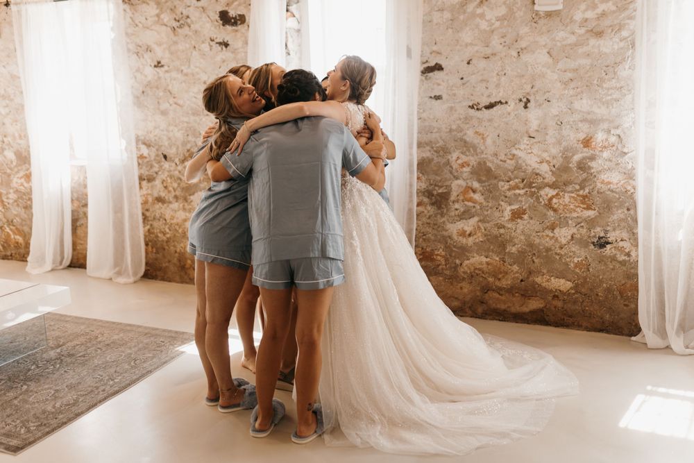 Bride in flowing lace wedding gown shares a warm group hug with bridesmaids in blue pajamas against rustic stone wall.