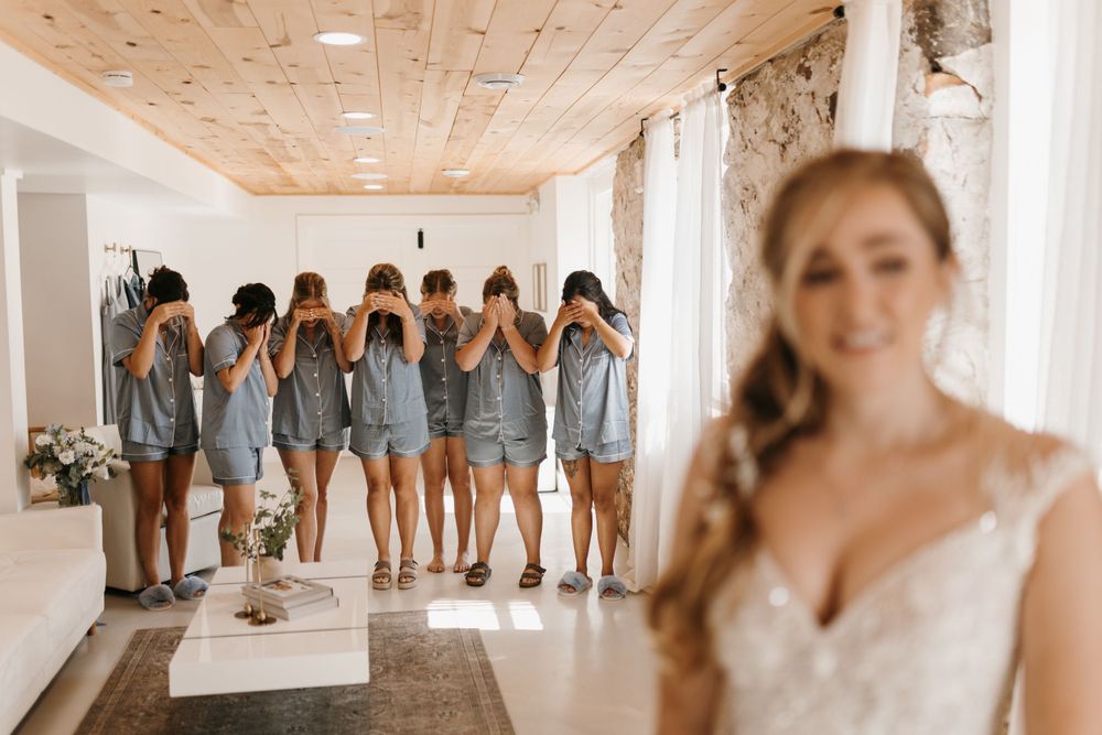 Smiling bride in lace gown poses while bridesmaids in matching blue pajamas cover their eyes during first look reveal.
