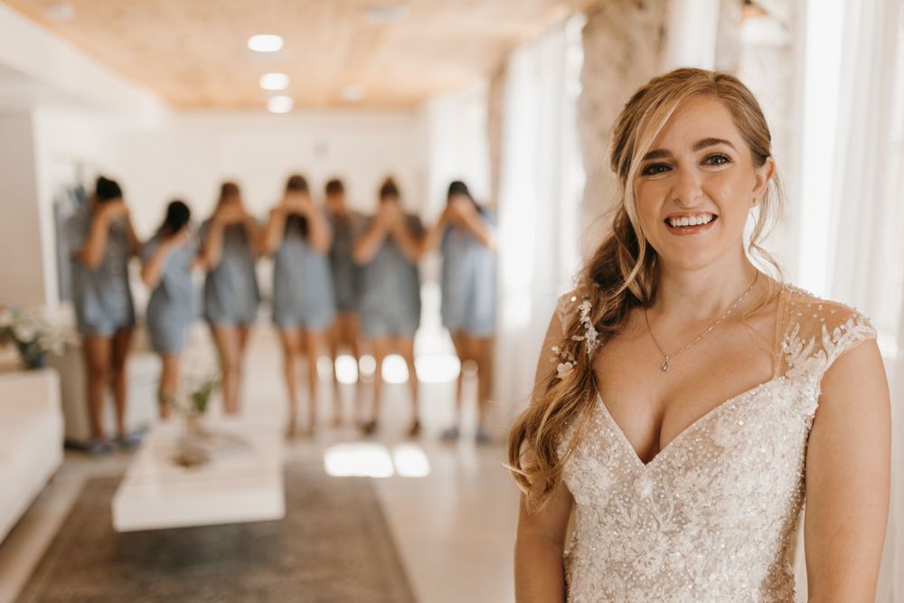Radiant bride in lace wedding dress smiles as bridesmaids in blue pajamas stand with backs turned in bright venue.