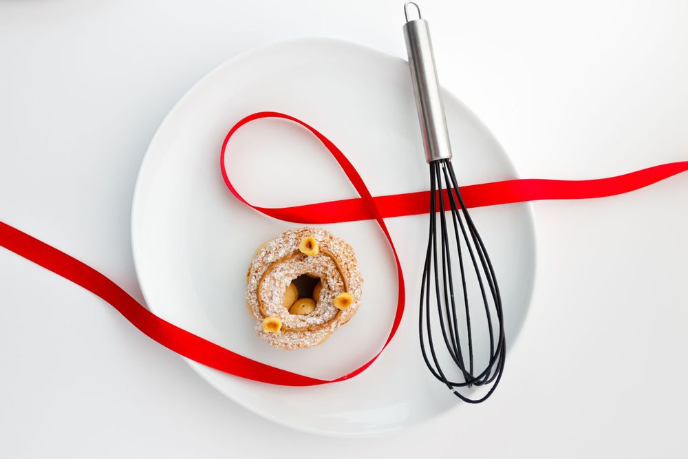 Top view of caramel pastry with red ribbon and whisk on white plate.