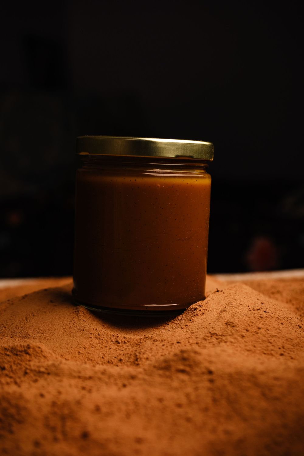 A glass jar of caramel spread sitting upright on a mound of cocoa powder.