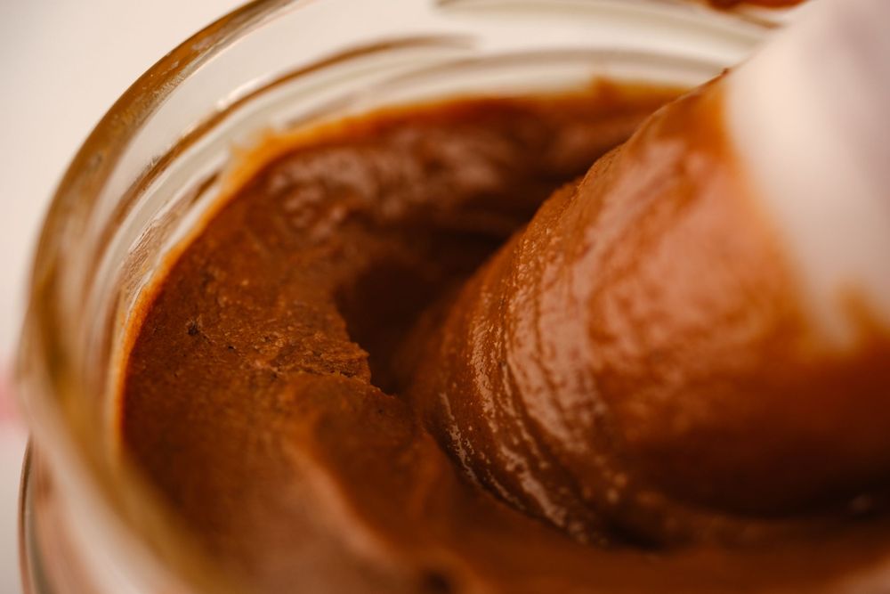 A close-up of a spoon scooping rich chocolate caramel spread from a glass jar.