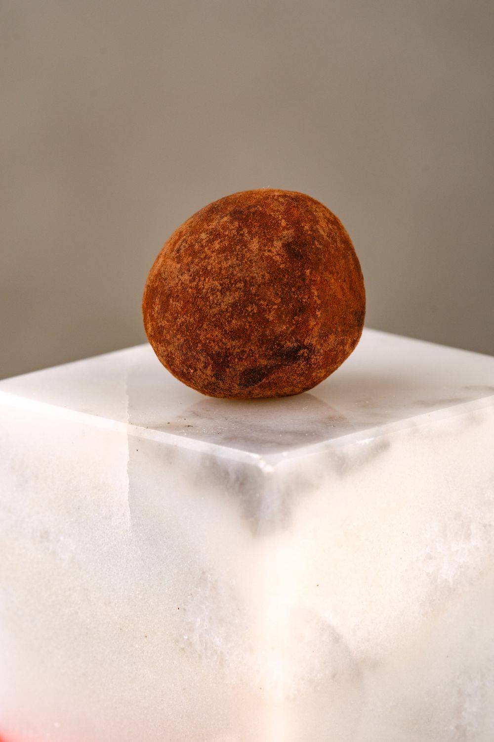 A single cocoa-dusted chocolate truffle resting on a white marble cube against a neutral background.